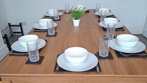 Una mesa de comedor dispuesta con platos, tazones, vasos y cubiertos, con una planta en maceta en el centro.