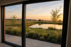 A view through a glass door to a garden with a pond, grass, and trees.