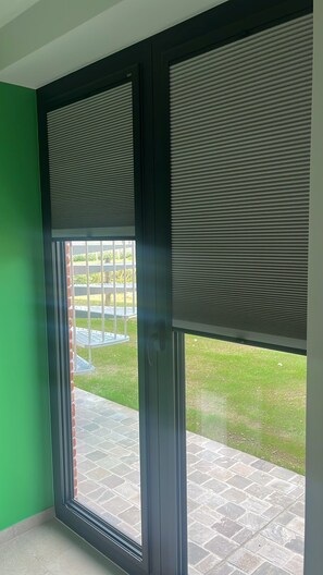 Een kamer met een grote schuifpui die uitkomt op een buitenruimte met een betegeld terras en een gazon.