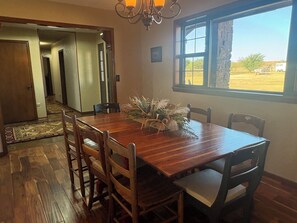 A dining area with a wooden table and chairs, a chandelier, a window with a view of the outdoors, and a door leading to another room.