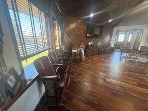 Una habitación con piso de madera, un televisor montado y una zona de estar con sillas.