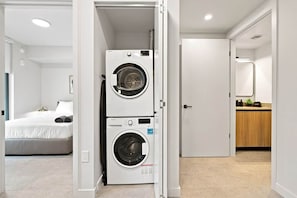 A laundry room with two stacked washing machines, a bed with white bedding, and a wooden cabinet in the background. - Downtown Miami 1006| Rooftop Pool Hot Tub & Gym (Miami)