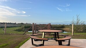 An outdoor seating area with a table and benches, surrounded by a fence and a view of a water body. - Den Kerselaer Group Holiday Home - Langemark       -   1st- FLOOR (Langemark-Poelkapelle)