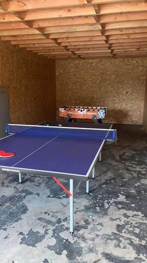 Une table de ping-pong avec un plateau bleu et des raquettes rouges, placée dans une pièce aux murs en bois et au sol en béton.