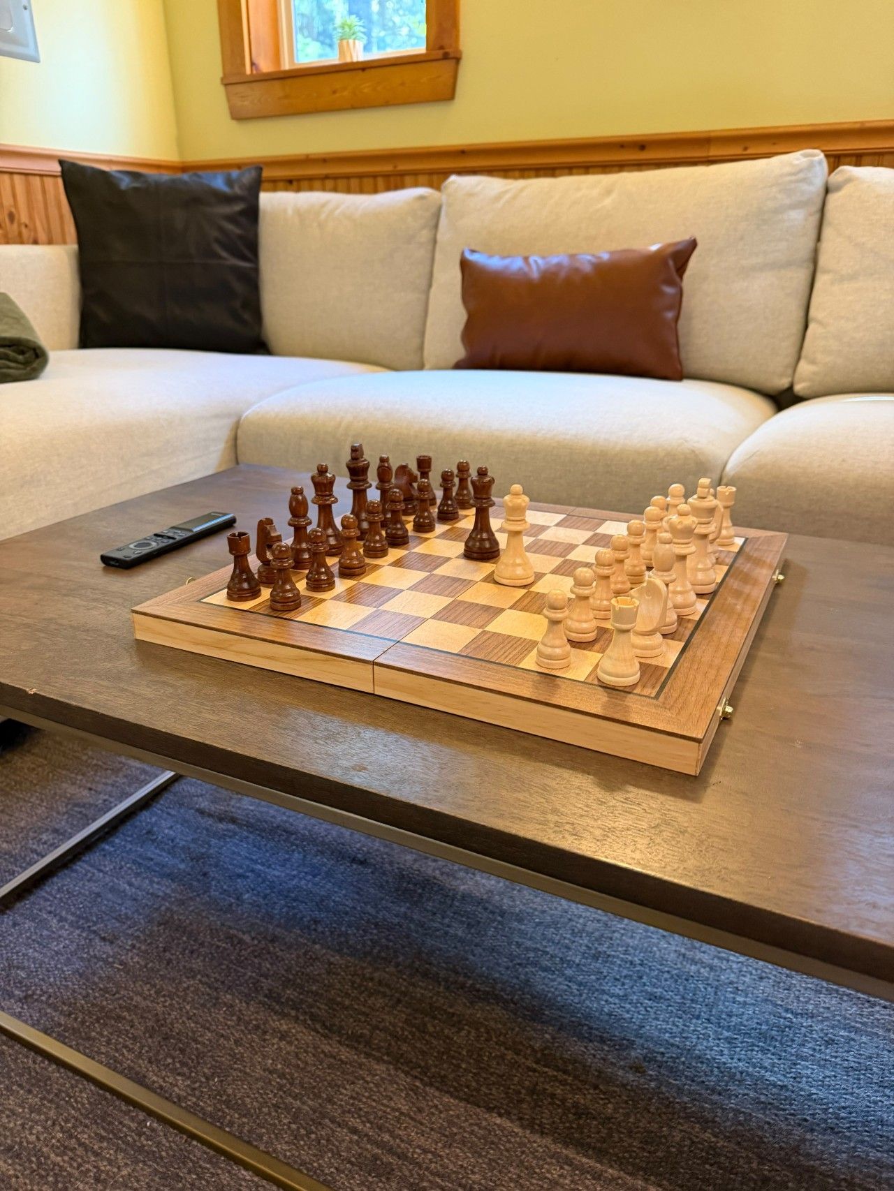 A chessboard on a wooden coffee table in a living room with a sofa and cushions in the background.