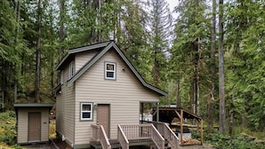 A two-story cabin with a small porch and a separate shed, surrounded by dense forest. - Your Cascade river retreat awaits (Marblemount)
