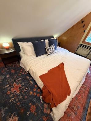 A bedroom with a bed, pillows, a blanket, a nightstand, a lamp, and a window.