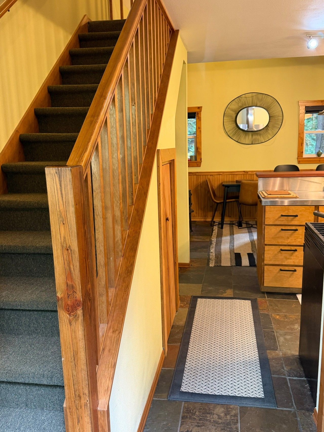 A staircase with wooden railings leading to a room with a dining area, a mirror, and a kitchen counter.