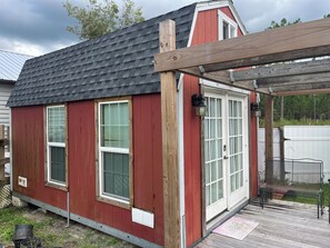 Un petit abri en bois peint en rouge, avec un toit incliné, une porte blanche et une fenêtre équipée de stores.