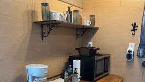 Et lille køkken med kaffemaskine, mikroovn og en hylde med glas.