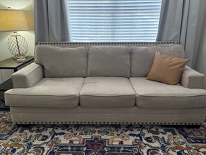 Ein graues Sofa mit einem einzelnen braunen Kissen, einem gemusterten Teppich und einer Lampe auf einem Beistelltisch.