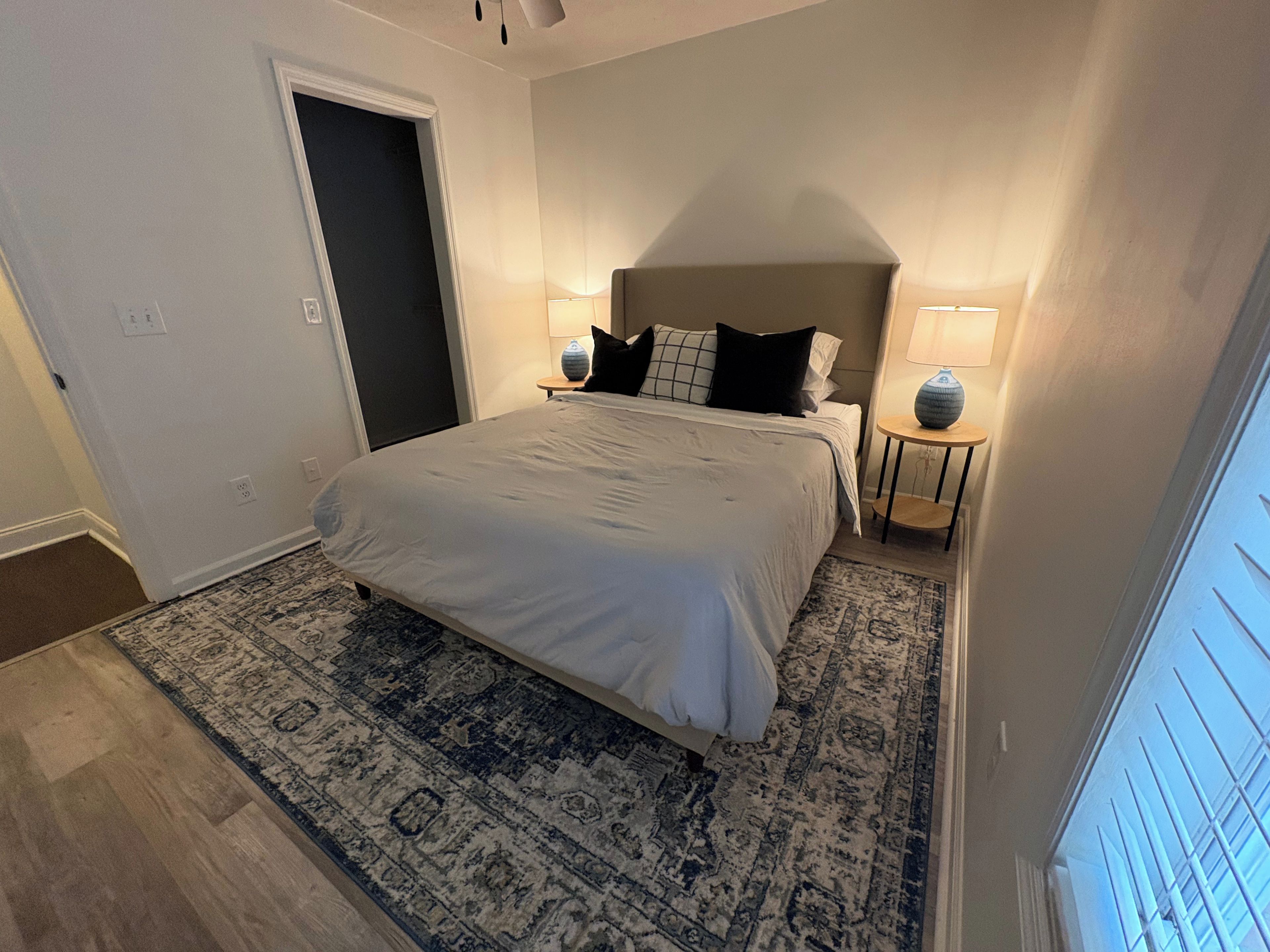 A bedroom with a large bed, a nightstand with a lamp, a door, and a patterned rug.