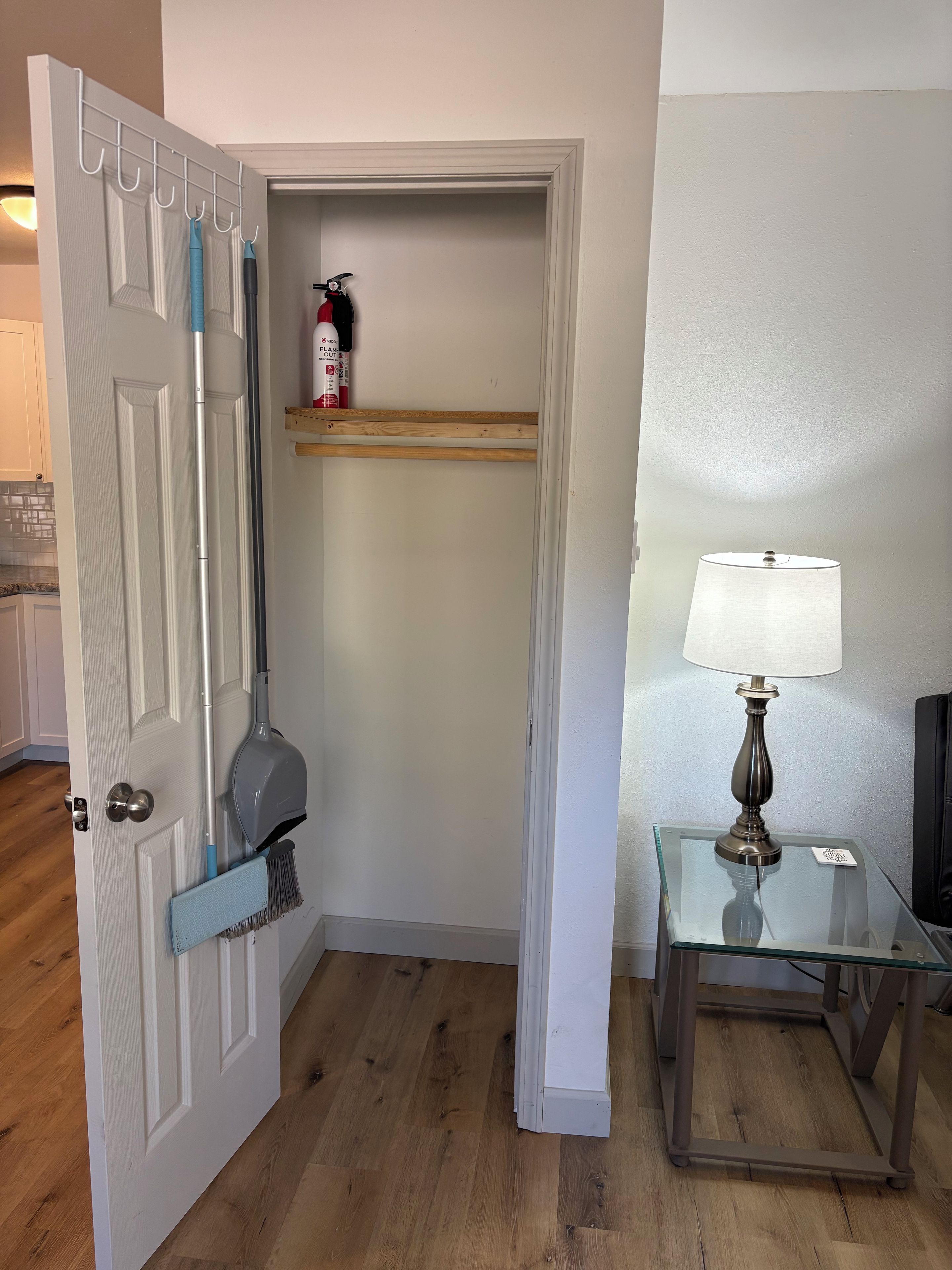 A room with a door, a mop, a broom, a shelf with a fire extinguisher, and a table with a lamp.