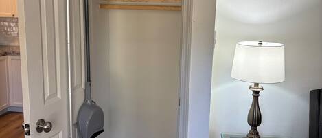 A room with a door, a mop, a broom, a shelf with a fire extinguisher, and a table with a lamp.