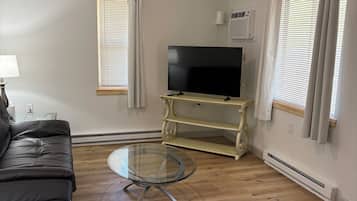 Un salon avec un canapé en cuir noir, une table basse en verre, un téléviseur à écran plat posé sur un meuble, un ventilateur de plafond et une fenêtre avec des stores.