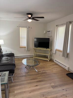A living room with a black leather sofa, a glass coffee table, a flat-screen TV on a stand, a ceiling fan, and a window with blinds. - Peaceful 1-bedroom apartment on large 2 acre lot. (Foster)