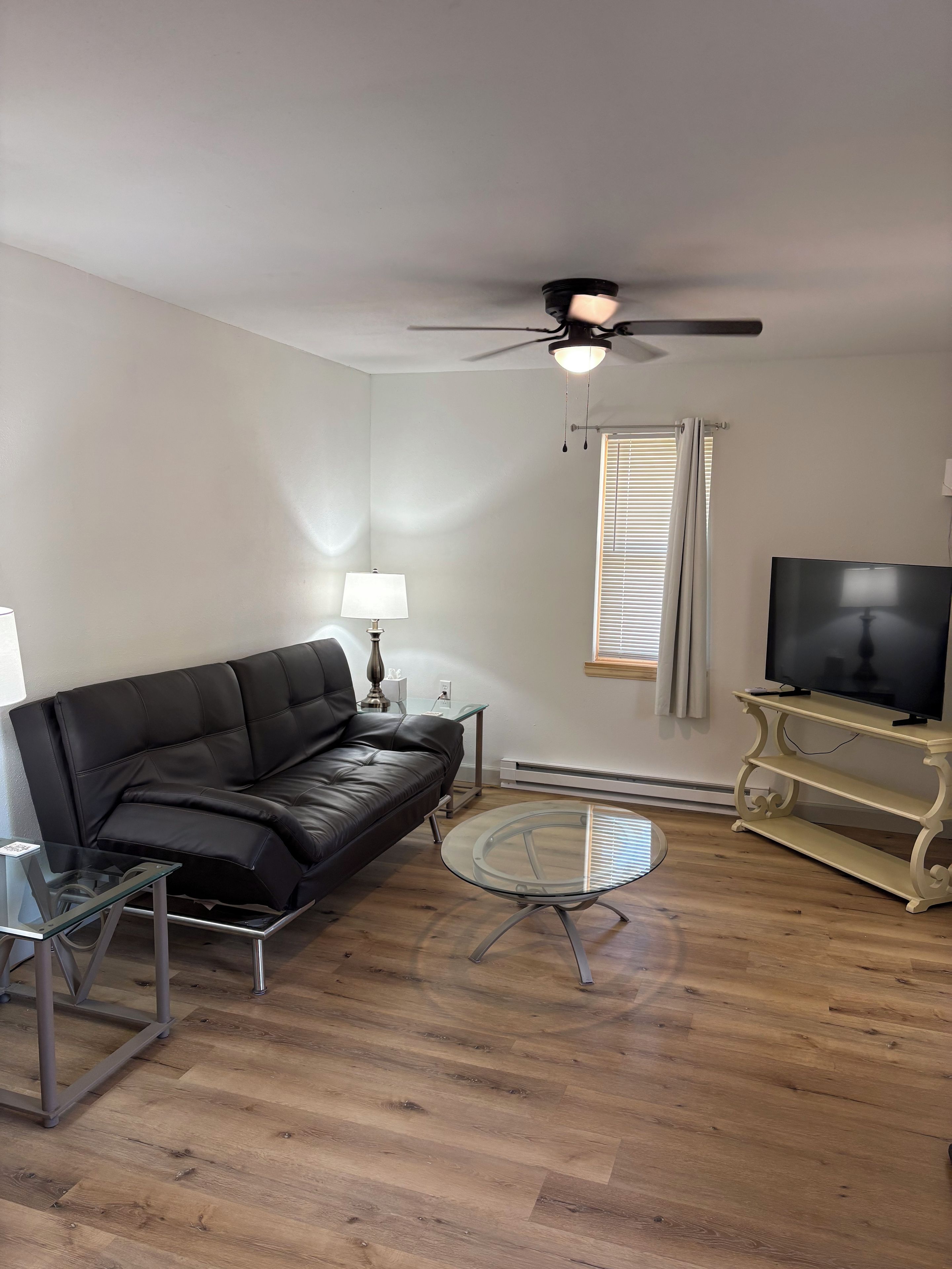 A living room with a black leather sofa, a glass coffee table, a TV on a stand, a ceiling fan, and a window with blinds.