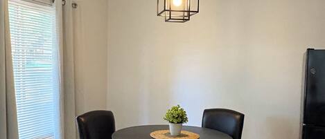 A dining area with a round table, two chairs, a hanging light fixture, a potted plant, and a trash bin.