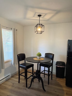 A dining area with a round table, two chairs, a hanging light fixture, a potted plant, and a trash bin. - Peaceful 1-bedroom apartment on large 2 acre lot. (Foster)