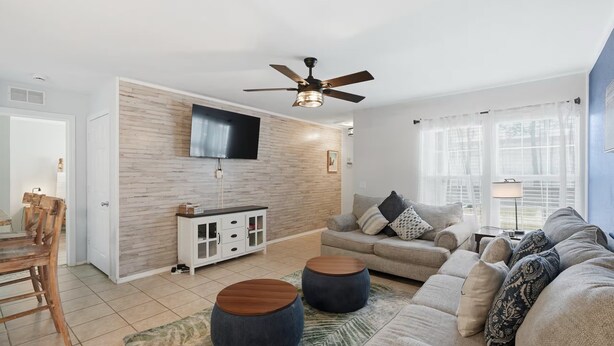 A living room with a TV mounted on a wooden wall, a ceiling fan, a sofa set, and a dining area with a wooden table and chairs. - Cape Cod Retreat 601-Pool & Playground-Sleeps 8 (Panama City Beach)