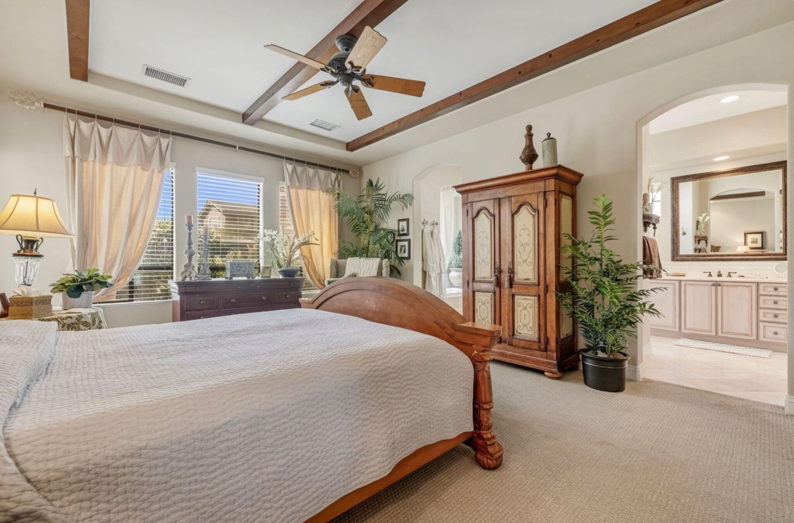 A bedroom with a large bed, a ceiling fan, a wooden dresser, and a bathroom with a mirror and cabinets.