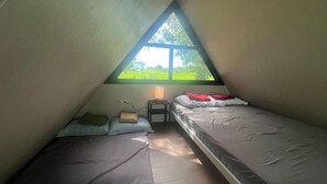 Una piccola camera da letto mansardata a forma di triangolo con due letti, un comodino e una finestra che offre vista sul verde.
