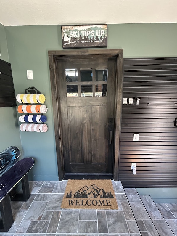A wooden door with a ski-themed sign above it, a welcome mat with a mountain design, and a set of striped towels hanging on the wall.