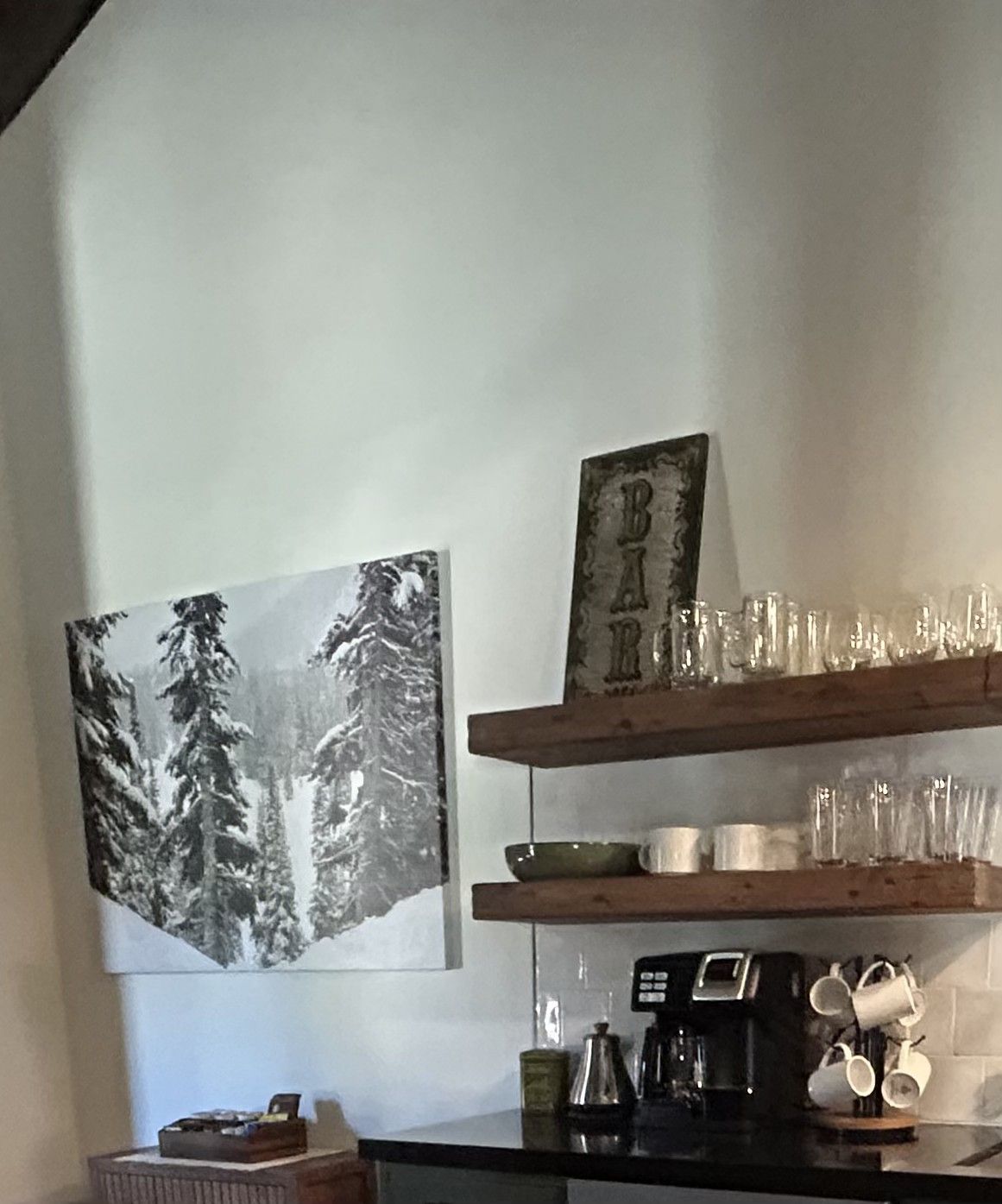 A kitchen with a coffee maker, a framed picture of trees, and shelves with glassware.