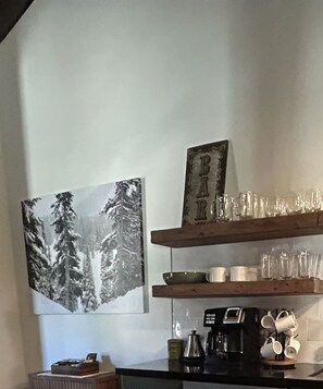 A kitchen with a coffee maker, a framed picture of trees, and shelves with glassware.