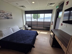 A hotel room with a bed, a chair, a desk, a window with a view of a lake, and a fire extinguisher. - Modern Deluxe Loft 103 (Peachland)