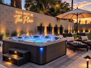 Een bubbelbad met een neonbord waarop staat 'THIS MUST BE The Place', in een achtertuin met lounge stoelen en een patio parasol.