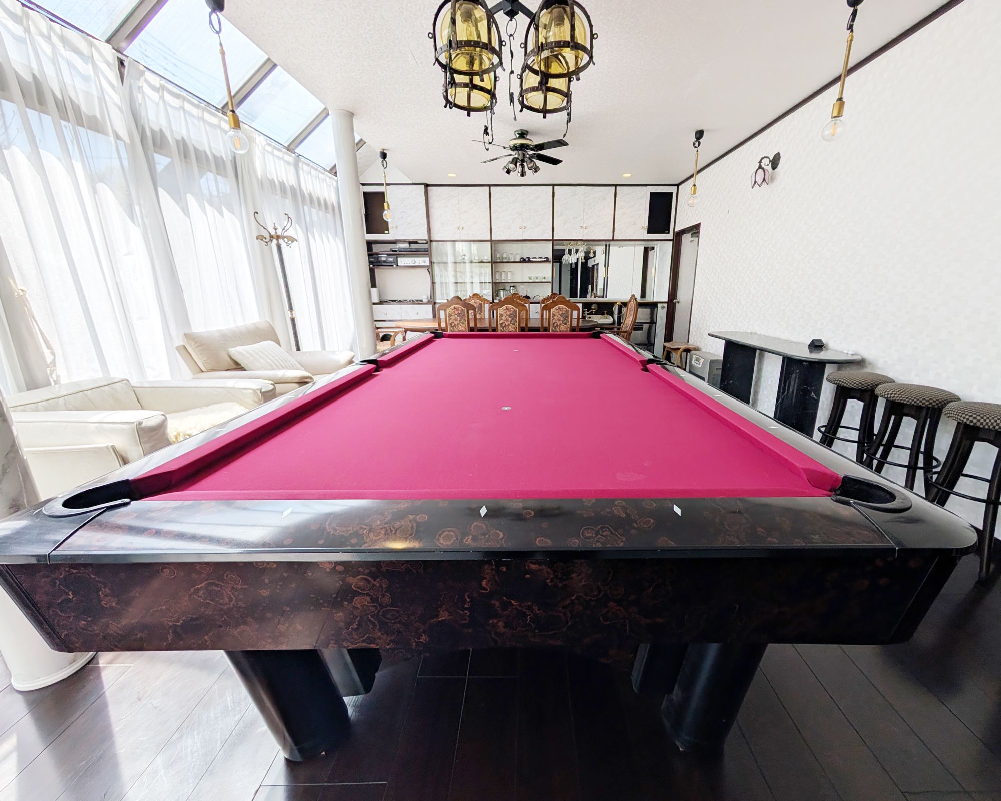 A pool table with a red felt surface, a ceiling fan, and a bar area with stools.