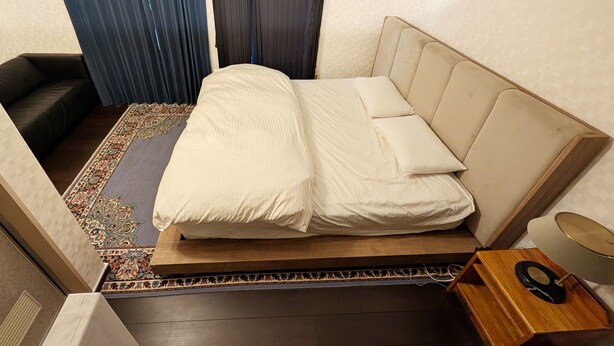 Quarto de hotel com cama, mesa de cabeceira pequena, luminária e janela com cortinas.