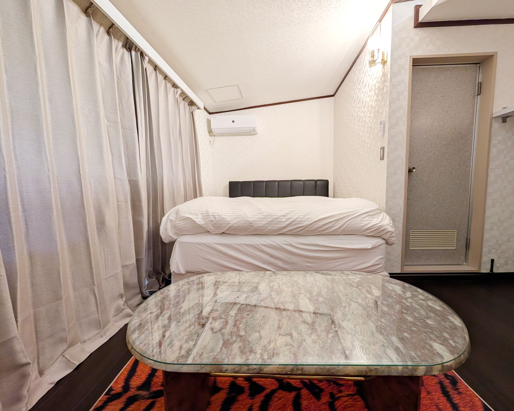 A hotel room with a bed, a glass-top table, a patterned rug, and a door.