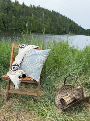 Een houten stoel met een gestreept kussen en een witte deken, geplaatst op het gras bij een meer met een bos op de achtergrond.