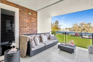 Ein Balkon mit einer grauen Rattan-Couch, einem kleinen Tisch mit Tablett und Blick auf einen Parkplatz und Bäume.