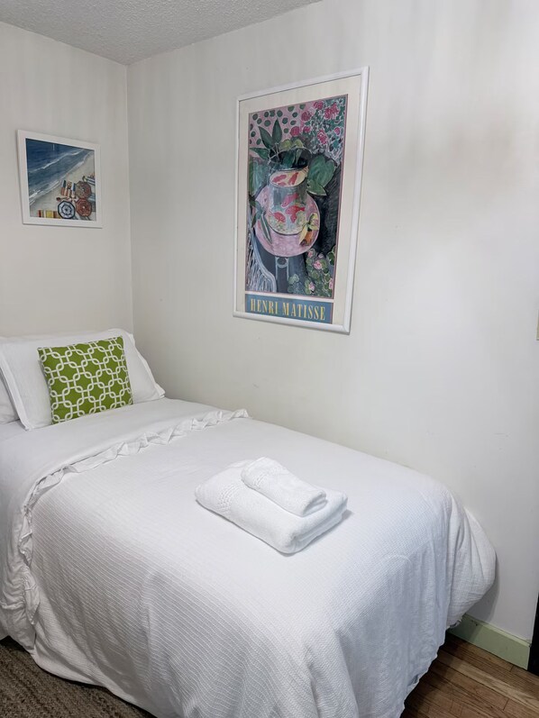 A neatly made bed with a folded towel, a green patterned pillow, and a framed artwork of Henri Matisse on the wall.