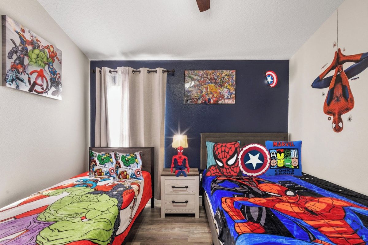 A child's bedroom with two beds, superhero-themed bedding, a Spider-Man pillow, and wall art.