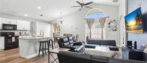 A modern kitchen with white cabinets, a central island, and a dining area with a black leather sofa and a coffee table.