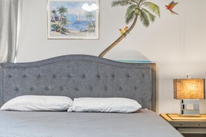 Ein Schlafzimmer mit einer gesteppten Kopfteile, einem Bett mit weißer Bettwäsche, einem gerahmten Gemälde von einer Strandansicht, einer Palmen Dekoration und einem Nachttisch mit einer Lampe.