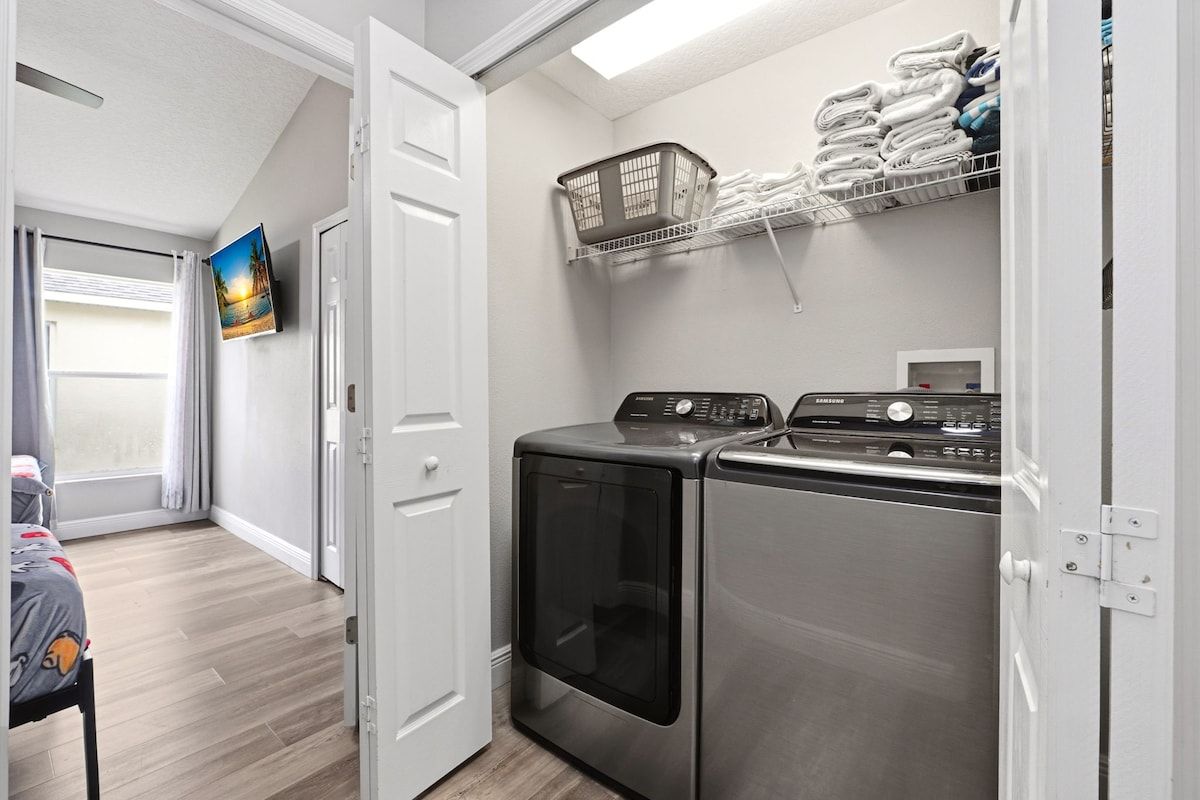 A laundry room with two stainless steel washers and dryers, a wall-mounted TV, and a shelf with folded towels.