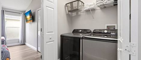 A laundry room with two stainless steel washers and dryers, a wall-mounted TV, and a shelf with folded towels.