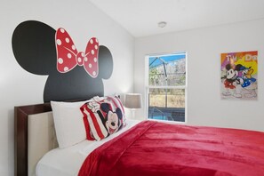 Un dormitorio con un mural grande de Mickey Mouse, una almohada con temática de Mickey, una colcha roja, una ventana con persianas y un cartel en la pared.