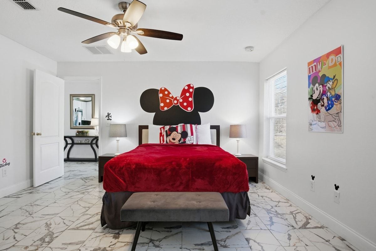 A bedroom with a bed, a ceiling fan, a Mickey Mouse head pillow, a red bedspread, and a marble floor.