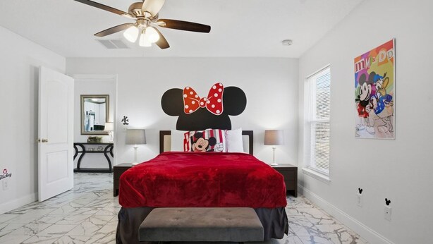 A bedroom with a bed, a ceiling fan, a Mickey Mouse head pillow, a red bedspread, and a marble floor.