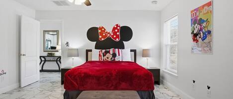 A bedroom with a bed, a ceiling fan, a Mickey Mouse head pillow, a red bedspread, and a marble floor.