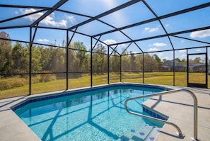A swimming pool with a metal frame and glass panels, surrounded by a metal fence, with a view of trees and houses in the background.