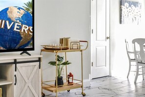 Una habitación con una televisión mostrando un globo terráqueo, un carrito dorado con una planta, y una puerta blanca.