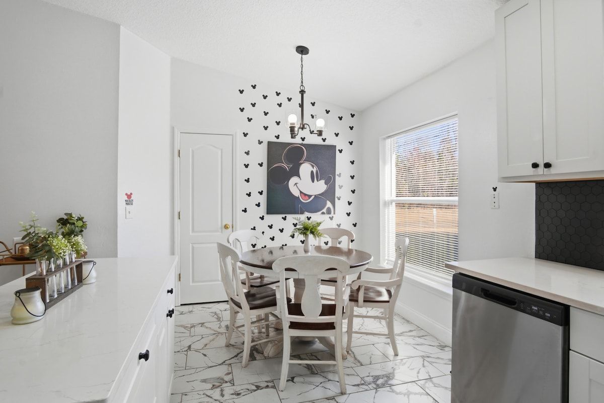 Une cuisine avec des meubles blancs, un sol en marbre, une table à manger avec des chaises, un lave-vaisselle et une fenêtre avec des stores.