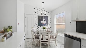Une cuisine avec des meubles blancs, un sol en marbre, une table à manger avec des chaises, un lave-vaisselle et une fenêtre avec des stores.